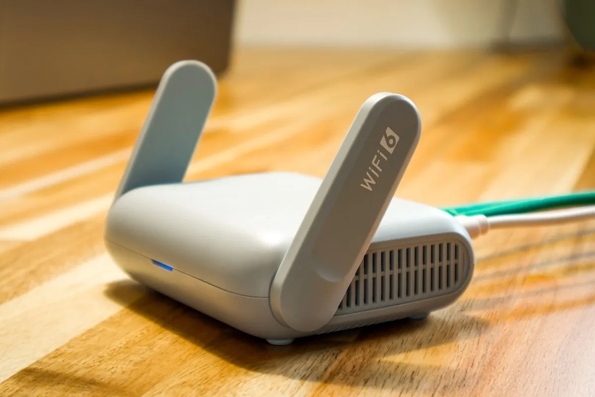 A compact grey travel router with two fold-up antennas on a wooden desk, with a laptop in the background.